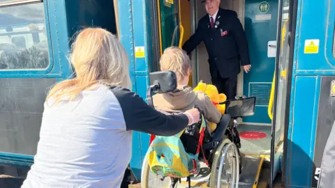 Emma Baugh/BBC A woman with long blonde hair, who is wearing a grey and black t-shirt, is pushing a wheelchair with a child in it up a ramp which is leading up to a blue train. There is a train worker standing at the door.