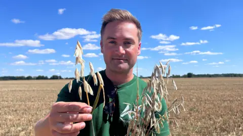 Tristan Ashby, standing in a field, holding onto some crops. A field is behind him with crops in it. He is wearing a green T-shirt, with sunglasses hooked on the front. He has short blonde hair. 