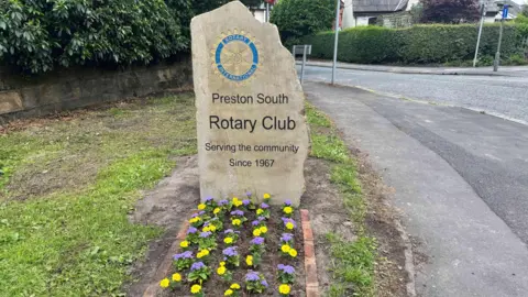 The newly installed stone marker on Liverpool Road, Penwortham, which reads Preston South Rotary Club - Serving the community since 1967. Purple and yellow flowers are planted at the foot of the stone marker.