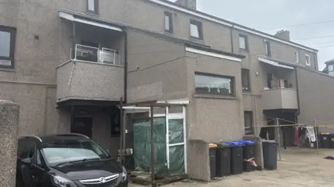Rear view of a grey block of flats. A black Vauxhall car is parked in the yard. Five grey wheelie bins are also visible as well as a washing line.