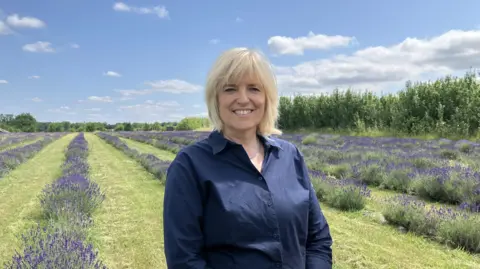 A woman in a dark blue shirt stood in front of rows of planted lavender, separated by grass. The sky is bright and blue with a few clouds.