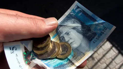 A hand holding a five pound note and several coins