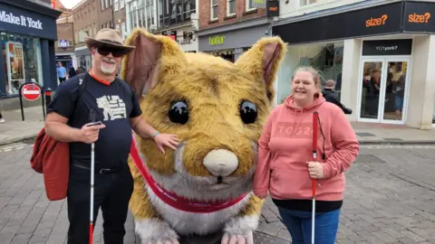 Supplied Julian Neale wearing glasses and holding a cane, next to a large hamster figure in a city centre, and a woman with a cane is stood on the right. 