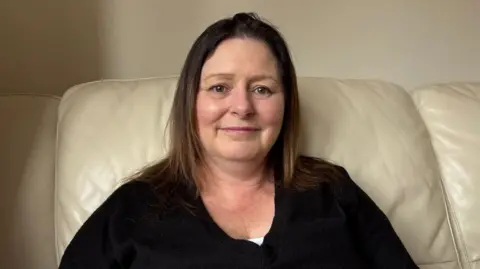 Martin Heath/BBC Brenda Brereton with long brown hair, smiling at the camera and wearing a brown sweater. She is sitting on a beige-coloured sofa