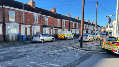 A police van and car are parked up.  The sun is shining down on the residential area of red brick terraced houses.