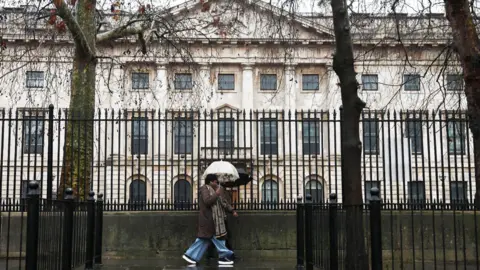 A woman holding an umbrella past black railings in front of the Royal Mint Court, the proposed site of the new Chinese embassy in London.