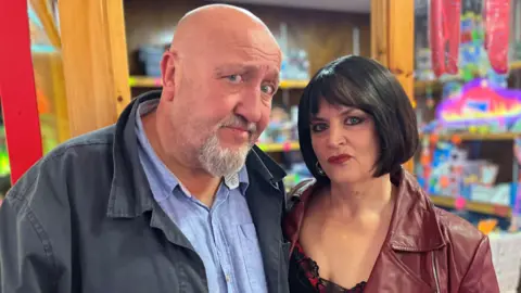 Ruth Jones, dressed in character as Nessa, standing next to Steve Speirs behind an amusements kiosk. Jones, on the right, wears a burgundy leather jacket, a black lace top and a black wig. She has red lisptick on and pouts at the camera with a stern look. Speirs, on the left, wears a blue linen shirt with a navy jacket. He looks at the camera with a confused expression.