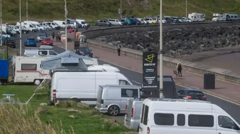 Motorhomes parked along a road on Scarborough’s North Bay with a wide pavement on the right hand side overlooking a beach with rocks in the background.