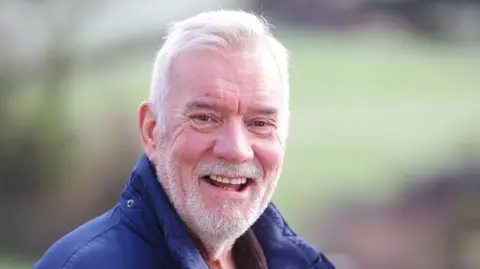 Brian Thompson A man smiles into the camera. He's wearing a navy coat and a brown top. He's outside. The background is blurred but he's outside.