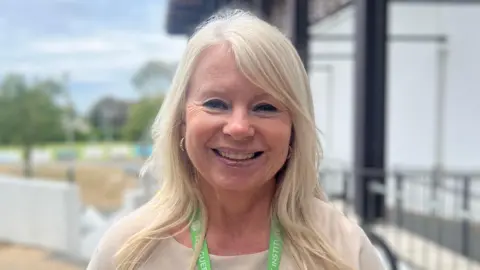 Katie Blompied - a woman with blonde hair, wearing a tan top, gold earrings and a green and white lanyard smiles at the camera.