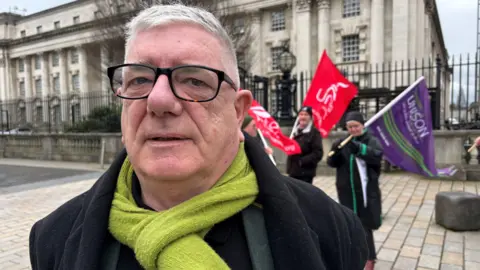 Gerry Murphy outside court. He is wearing a black jacket, green scarf and glasses. He has grey hair. Behind him are campaigners with red and purple flags. 