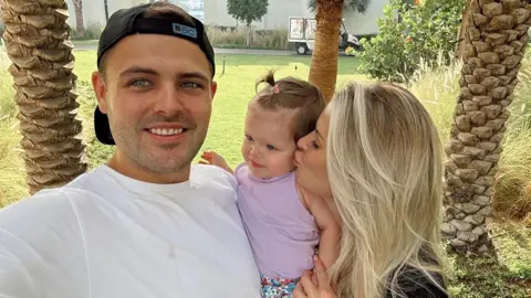 Robyn Baker Robyn's partner is on the left. He has brown hair underneath a baseball cap, and is wearing a white top. In the middle is their daughter, and on the right is Robyn kissing their daughter.