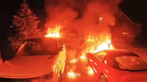 A pitch black night with a white Range Rover on the left of a drive next to a Mitsubishi. Both are covered in flames and the drive itself is on fire. A tree can be seen in the background lit by the orange glow.