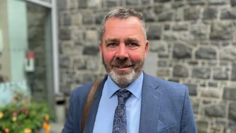 A man in a blue suit smiles at the camera. He is wearing a blazer, blue shirt and paisley-patterned tie. He is standing outdoors in front of a stone wall. 