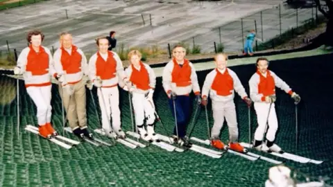 Kendal Snowsports Club A row of skiiers wearing red and white ski clothing and ski boots. The image quality is quite blurry because it was taken in the 1980s. They are standing lined up with skis, poles on green dry slope matting. 