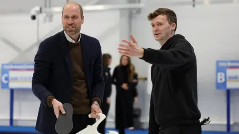 PA Media Prince William standing on a curling rink with equipment in both of his hands while a staff member stands next to him with his hand out demonstrating. 