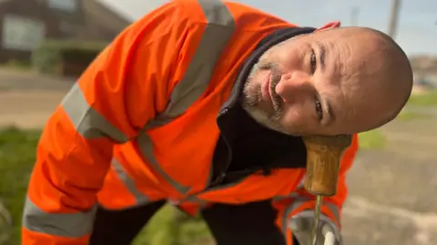 Fiona Irving / BBC A man with a shaved head and a beard and wearing a high-vis orange jacket is looking at the camera. He has his ear to wooden cylinder-shaped object which is attached to a metal rod.