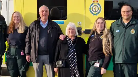 Five people smiling, standing in line, facing the camera, with an ambulance behind them. A man and woman with grey hair are in the middle, and are flanked by two women and a man, who are all in ambulance service uniform.