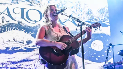 Getty Images The image shows a person performing on stage, playing an acoustic guitar and singing into a microphone. Behind them is an artistic backdrop featuring various illustrations.