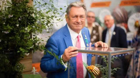 A man, Alan Titchmarsh, wearing a blue suit with a pink tie. He is stood in front of a glass container, holding a large model of a beetle. The beetle has cream and black stripes on its body and orange and black spots on its head.