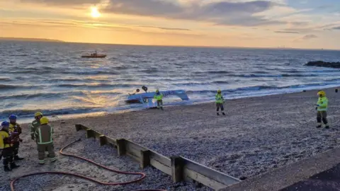Hampshire & Isle of Wight Fire and Rescue Service A light aircraft is upside down in shallow water by the beach with emergency services personnel standing nearby