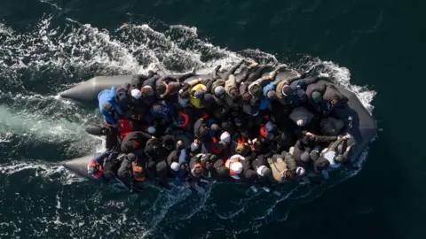 Dozens of people are crammed onto a small black dinghy, which leaves white trails of foam behind it in the sea