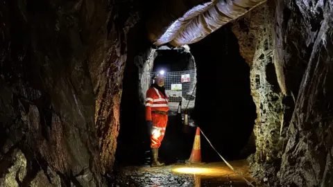 A miner dressed in orange and wearing a hard hat looks at the camera while standing in an underground mine. There is an orange and white cone beside him and his helmet lamp is reflecting off a puddle on the ground beside him.
