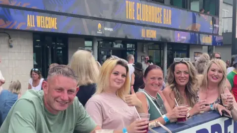 A man on the left and four women are holding drinks and looking at the camera. Other people are visible in the background at the bar.
