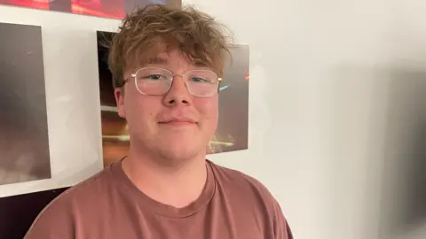 A teenage boy has long brown hair and silver glasses. He is smiling and is wearing a dusky terracotta colour t-shirt