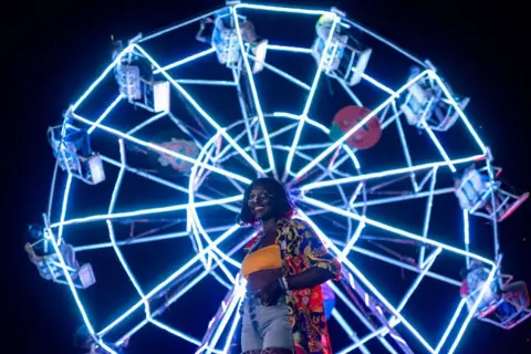 Badru Katumba / AFP A woman stands in front of a ferris wheel that is brightly lit up at the Nyege Nyege festival in Uganda - Saturday 16 November 2024