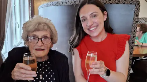 Hannah Kent Two people sit at a restaurant table holding glasses of sparkling wine. One wears a red ruffled top, and has long dark brown hair Hannah. the other, Joan, is elderly and has shorter blowdried hair and glasses. Joan wears a black cardigan over a polka-dot top. Behind them is a tall booth with patterned fabric and round mirrors on the wall.