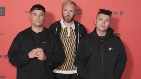 Mo Chara, left, is wearing a black sweatshirt and looking directly at the camera. He had short brown hair. DJ Provai, middle, is wearing a yellow and green patterned polo top and a black zipper jacket. He is looking directly at the camera and has a beard and ginger hair. Moglai Bap, is wearing a black zip up coat. He is wearing sunglasses on his head and has short brown hair.