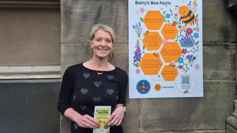 Dr Kate Stevens - a woman wearing a black top with white hearts on it, standing outside next to a large brightly coloured sign reading Barry's Bee Facts
