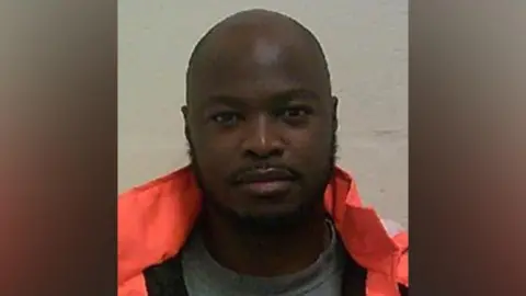 A bald man with a beard and moustache in a police mugshot. He is wearing a bright orange coat.