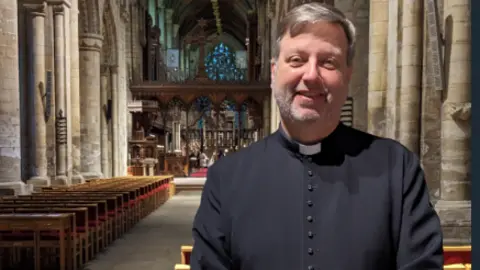 James Graham/BBC A smiling man wearing a black gown with a white clerical collar round his neck. He is stood on a church setting with chairs and ornate pillars in the background. 