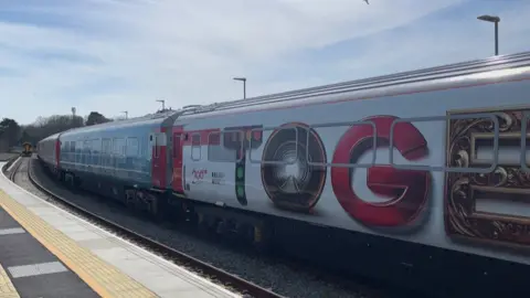 A train comprising four carriages with the words Railway 200 and Inspiration Tour is pulled into Newquay station under a blue sky. There are two railway lines - the one not in use by the exhibit train has one depaerting away from Newquay. On the exhibit train - the carriages are brightly coloured and they each have a different exhibition inside them. They are called Railway Firsts (history), Wonderlab on Wheels (interactive engineering), Your Railway Future (careers/innovation), and a Partner Zone.