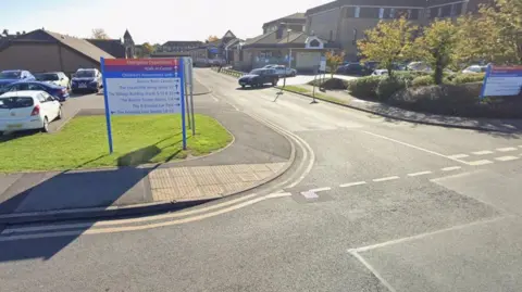 A hospital car park. A sign indicating the way to different departments, parked cars and hospital buildings can all be seen.