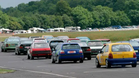 Getty Images A view of the track with a number of usual road cars adapted for racing on circuits - old hatchbacks and small saloons in a variety of colours.. About to set off racing. In the distance, caravans and cars parked.