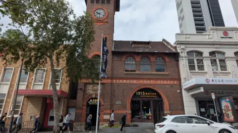 People walk along the pavement in front of a clock tower at Burwood, Reid, next to a Bank of China branch office
