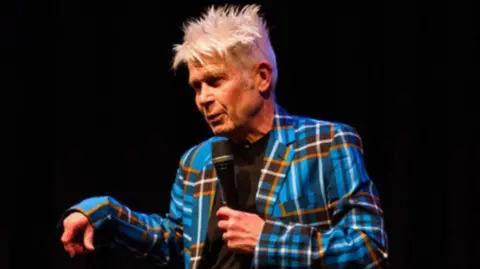 Vladimir McTavish on stage. He is wearing a blue, black, white and orange tartan suit and is holding a microphone in his left hand.