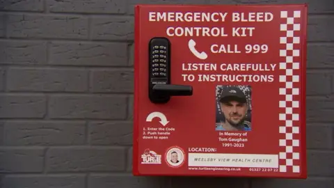 A red bleed kit box. It includes a black and silver key code, a photo of Tom Gaughan and white writing that reads: "Emergency bleed control kit. Call 999. Listen carefully to instructions. In memory of Ton Gaughan, 1991-2023."