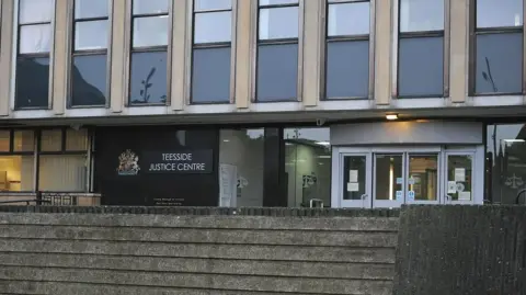 The exterior of the grey, concrete court building. A floor-to-ceiling black sign next to the glass doors reads: "Teesside Justice Centre". The second storey of the building is made of alternating blacked out windows and light cream concrete. The wall in front of the reception is made of grimy grey stone.