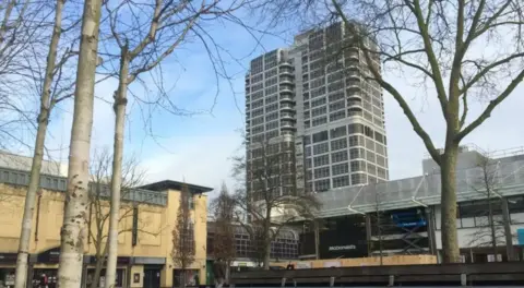 A grey building with rounded edges which towers over nearby buildings. There is a shopping centre in the foreground. There are trees either side of the image.