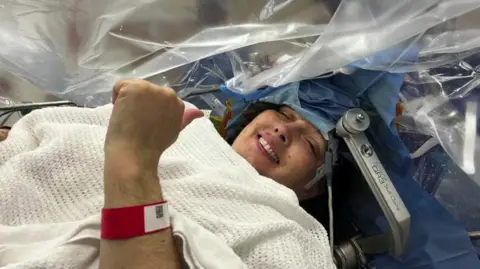 Lizzie Pitt A woman lies awake during a surgical procedure, her head secured in a frame and covered with sterile drapes, giving a thumbs‑up to the medical team.