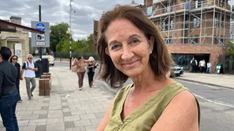 Tom Jackson/BBC Head and shoulders image of Heather Boyd. Heather has shoulder-length brunette hair. She has a light green sleeveless top and is stood on a footpath with a building under construction behind.