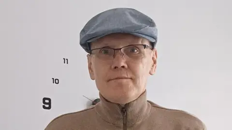 Robert Best is wearing glasses, a light blue, flat cap, and a tan zip-up jumper. He is standing in front of a white wall with a large minimalist clock showing the numbers 9, 10, and 11.