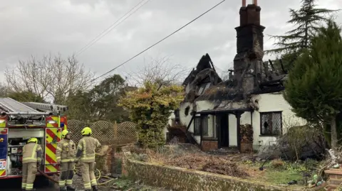 Luke Deal/BBC Firefighters stand near their fire engine outside the cottage. They wear firefighting uniforms and safety hats.