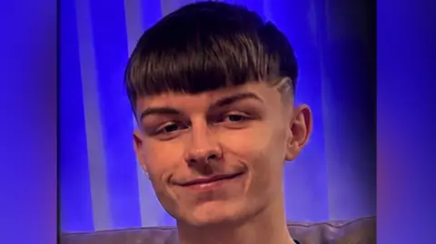 A teenage boy smiling at the camera. He has short brown hair and a black t-shirt on.