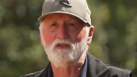 Nigel has a white beard and is wearing a grey cap and dark jacket.