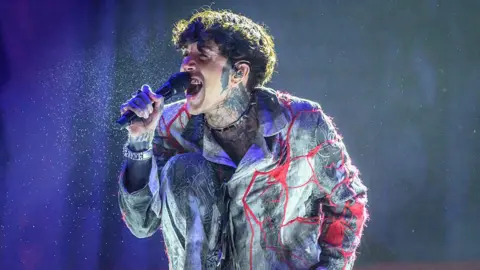 Oli Sykes performing onstage during Leeds Festival at Bramham Park in 2025. He has curly brown hair. He is singing into a mic wearing a red and grey shirt-jacket. He has tattoos on his hands wrists, neck and face. 